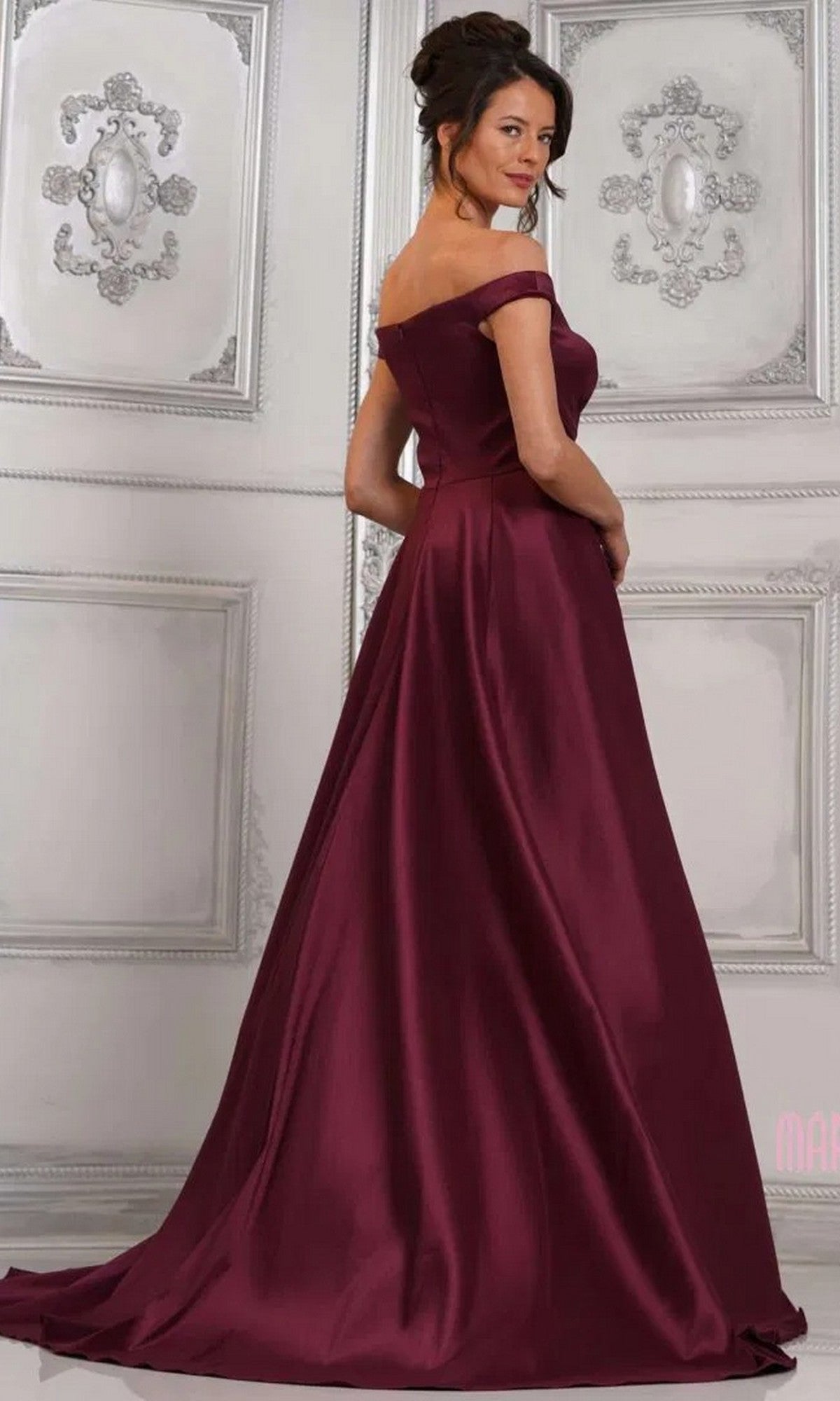 Wearing the Marsoni MV1310, a long formal off-the-shoulder mermaid gown, a woman stands in an elegantly decorated room, gazing over her shoulder.